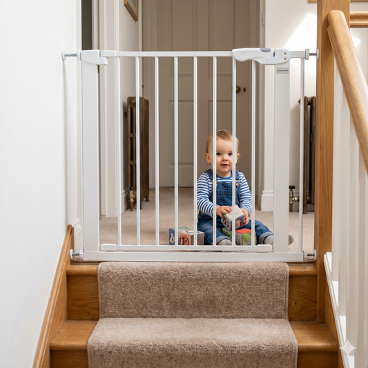 A toddler in blue overalls sits on a carpeted hallway, playing with soft blocks behind a securely closed white screw-fit baby gate at the top of a wooden staircase, representing safe installation.
